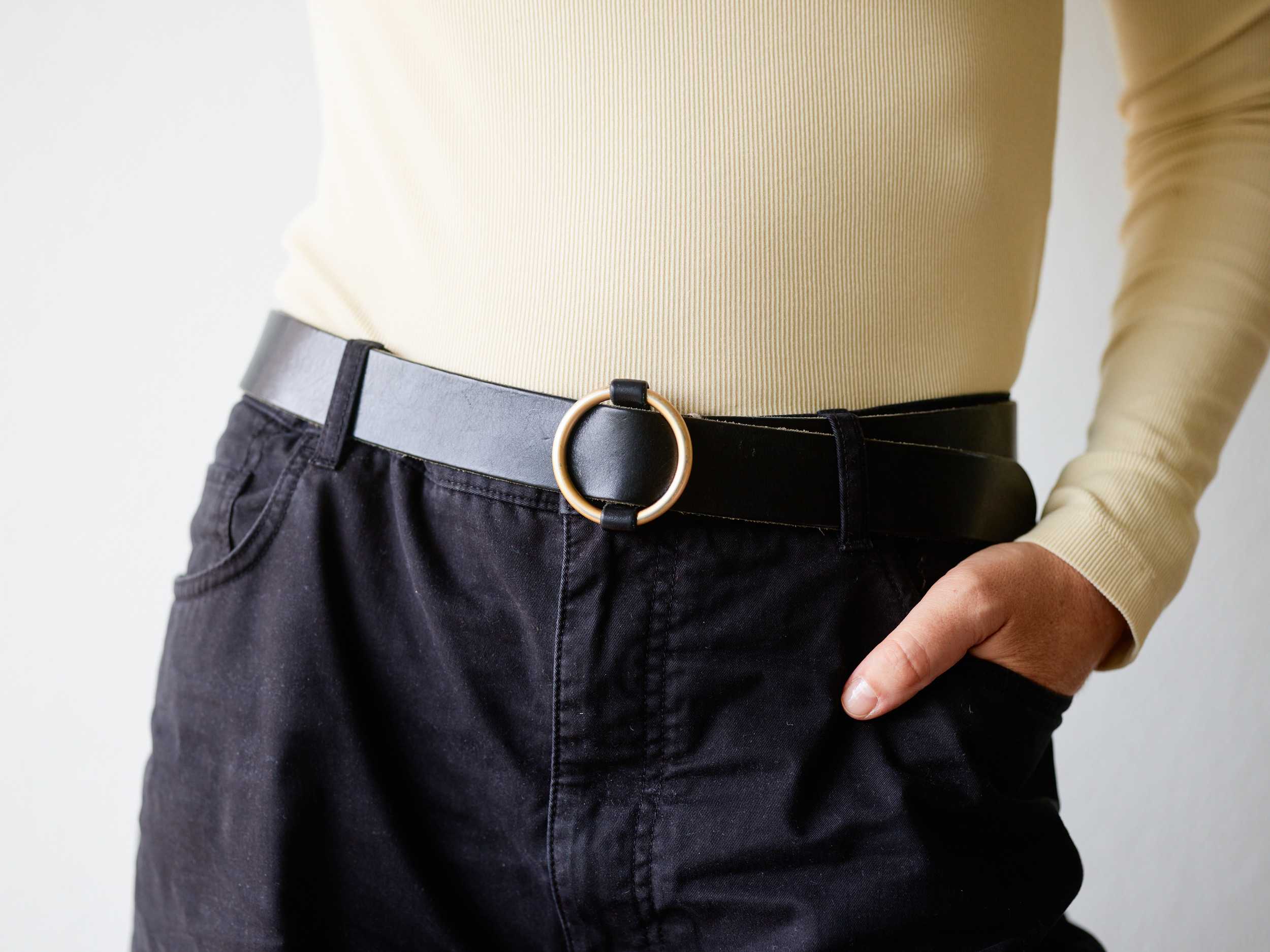 Person wearing a beige ribbed long-sleeve top tucked into black pants with a black belt featuring a round gold buckle